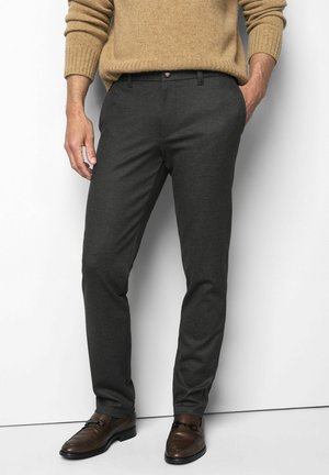 Mann trägt dunkelgraue Slim-Fit-Hose, braune Lederschuhe und einen beigen Strickpullover mit einer Hand in der Tasche.
