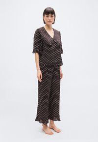 Femme debout pieds nus portant un chemisier assorti à pois marron foncé avec col à volants et un pantalon à jambes larges avec ourlets à volants.