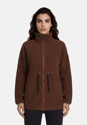 Vrouw die naar voren kijkt en een bruine fleecejas met rits, hoge kraag en zwarte trekkoorden draagt over een zwarte broek.