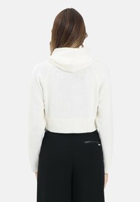 Maglione bianco corto con cappuccio, trama lavorata a maglia, orlo e polsini a coste, mostrato di spalle su uno sfondo semplice.