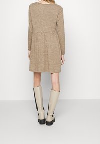 Braunes langärmliges Kleid mit gerafftem Taillenbereich, aus strukturiertem Material gefertigt. Kombiniert mit kniehohen beigen Stiefeln mit schwarzem Akzent.