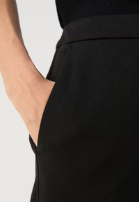 Main reposant dans la poche avant d'un pantalon noir taille haute sur un fond clair.