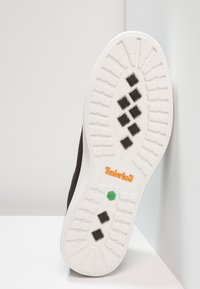 Baskets en daim noir avec une semelle en caoutchouc blanche. Elles présentent un motif de grip texturé, un logo « Timberland » orange et un détail accent vert.