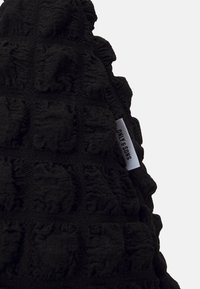 Tissu noir texturé avec des motifs carrés en relief et une étiquette blanche indiquant "SINS & ONLY" cousue dans la couture.