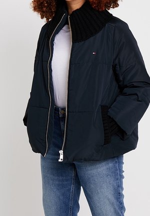 Femme portant une veste matelassée bleu marine avec col et poignets en tricot noir, chemise blanche et jean bleu, mains dans les poches de la veste.
