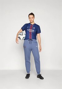 Blaues Fußballtrikot mit roten und weißen Akzenten, Logo und Schriftzug. Graue Jogginghose mit Kordelzug und Logo. Schwarze Fußballschuhe. Hält einen Fußball.
