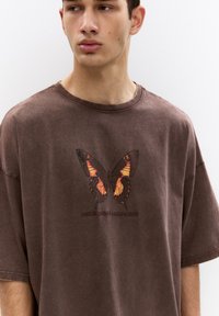 La maglietta oversize marrone presenta un design dettagliato di una farfalla in arancione e nero, con il testo "NATURE SERVICE LOS ANGELES" sottostante.