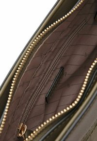 Intérieur d'un sac vert olive avec une doublure marron matelassée, comportant une poche zippée et une petite étiquette, fermée par une fermeture éclair dorée.