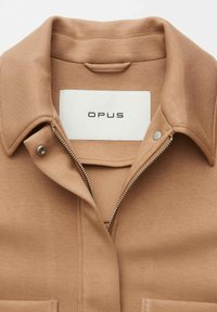 Manteau marron à texture douce, doté d'un col pointu, d'une fermeture éclair argentée et d'une étiquette blanche avec l'inscription "OPUS" en lettres noires.