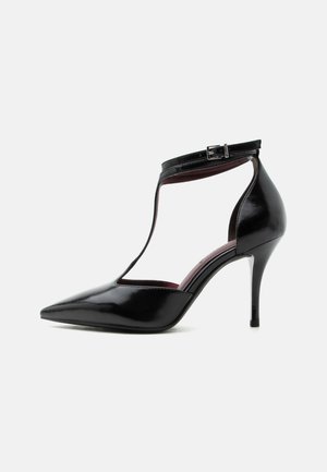 Scarpa con tacco alto in pelle verniciata nera con punta appuntita, cinturino a T e chiusura con fibbia intorno alla caviglia.