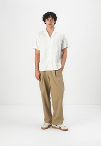 Short-sleeve, white shirt with textured leaf pattern, paired with loose beige trousers and cream sneakers with dark accents.