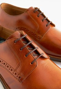 Smeđe kožne cipele s glatkom teksturom, s brogue detaljima, okruglim vrhom i smešnim vezicama na smešnom ulošku.