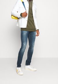 Giacca bianca sopra un felpa verde oliva, jeans skinny blu strappati e sneaker bianche con dettagli gialli. Modello che porta uno zaino giallo.