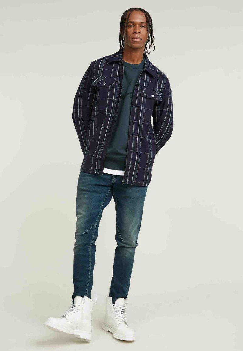 Marineblauwe geblokte shirtjas met twee borstzakken, gedragen over een donkergroen sweatshirt, gecombineerd met blauwe jeans en witte hoge sneakers.