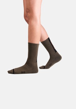 Jambes portant des chaussettes côtelées vert olive avec des accents noirs au talon et aux orteils, taille 39-42, sur un fond clair uni.