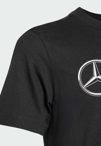 Černé tričko vyrobené z bavlny s kulatým výstřihem, se zřetelně vyobrazeným bílým logem Mercedes-Benz na hrudi.