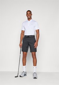 Camisa polo branca de mangas curtas e colarinho, combinada com calções pretos. Ténis cinzentos e meias brancas. Taco de golfe segurado na mão.