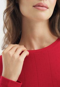 Red ribbed top with a wide neckline, featuring small texture patterns. A hand gently holds the fabric near the collar, showcasing the material's softness.