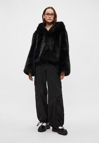 Juoda dirbtinio kailio striukė su plačiomis rankovėmis ir gaubtu, derinama su laisvomis juodomis kargo kelnėmis ir masyviomis juodomis platforminiais batais.