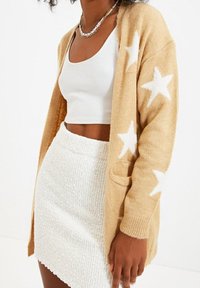 Beige stickad kofta med vita stjärnmönster, öppen fram, fickor och ribbade ärmslut. Bärs över en vit crop top och en texturerad vit kjol.