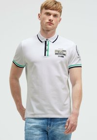 Weißes Poloshirt mit schwarzen und grünen Akzenten, drei Knöpfen und Patches auf der Vorderseite und dem Ärmel. Kurze Ärmel und klassischer Schnitt. Lässiger Stil.