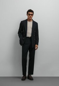 Blazer croisé bleu marine foncé, chemise beige à col rond, pantalon sur mesure, chaussures marron, lunettes rectangulaires ; textures lisses et lignes épurées.