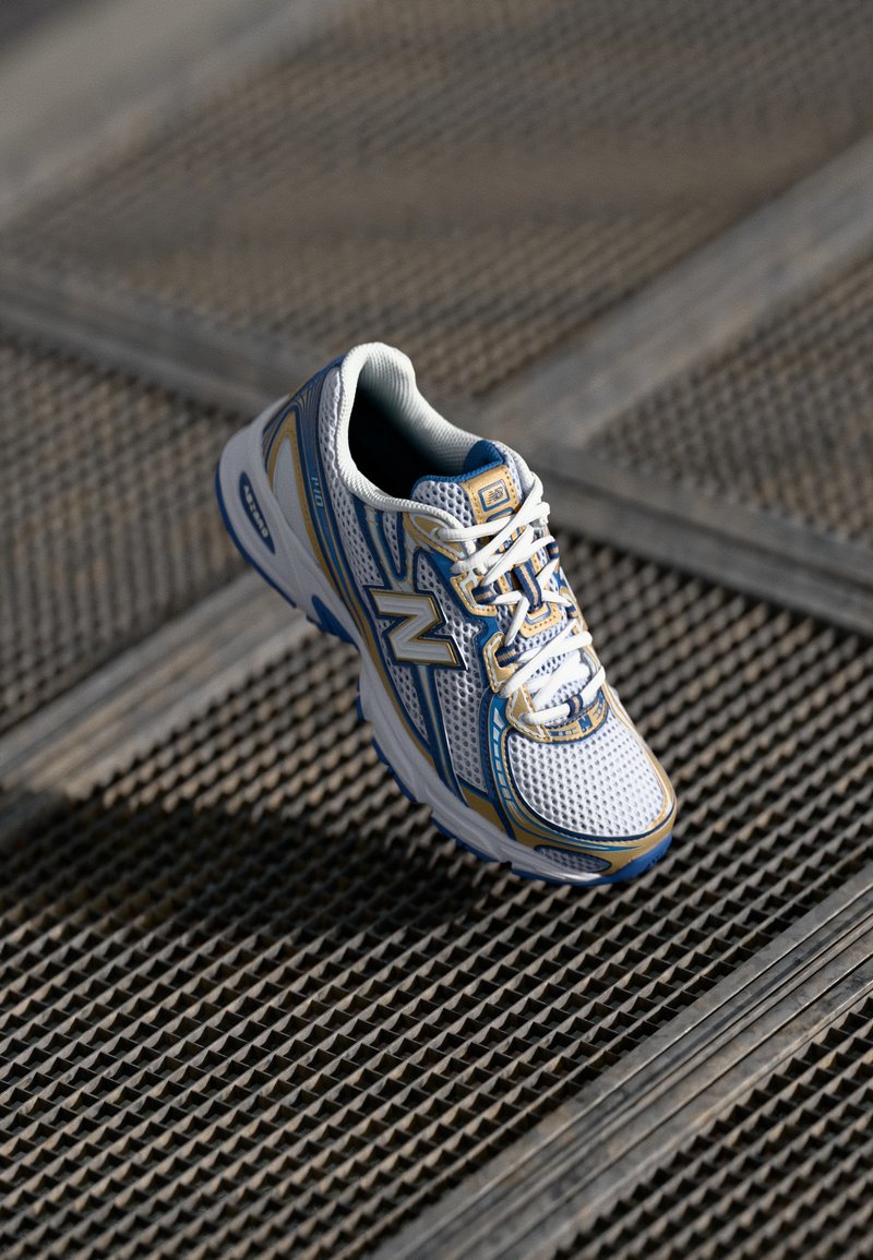Zapatilla deportiva blanca con acentos en azul y dorado. Presenta una parte superior de malla, cordones y una suela texturizada. Diseño ligero adecuado para deportes.