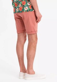 Helle rosa Baumwollshorts mit umgekrempelten Säumen, kombiniert mit weißen Slipper-Schuhen; im Hintergrund ist ein blumig gemustertes Hemd zu sehen.