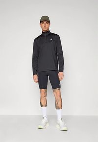 Bluză neagră cu mâneci lungi și fermoar, cu logo, combinată cu pantaloni scurți închiși la culoare. Modelul poartă o șapcă, șosete albe și pantofi de alergare cu talpă deschisă la culoare.