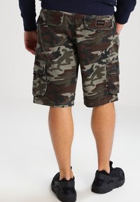 Tummanvihreät ja ruskeat maastohousut, joissa on useita sivutaskuja ja ommeltu etiketti. Materiaali on kestävä ja teksturoitu.