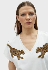 T-shirt bianca con scollatura a V che presenta due applicazioni di leopardo in perline oro e nero su ciascun spalla. Orecchini geometrici dorati.