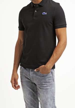 Schwarzes Poloshirt aus Baumwolle mit Kragen, kurzen Ärmeln und einem blauen Lacoste-Logo auf der Brust. Getragen zu hellgrauen Jeans aus Denim.