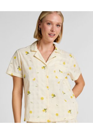 Blonde Frau trägt eine cremefarbene Kurzarm-Bluse mit gelbem Zitronenmuster und kleinem vorderem Taschenaufschlag, lächelt vor einfachem Hintergrund.