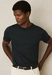 Tricou negru din bumbac, cu guler rotund și mâneci scurte, purtat cu pantaloni albi și o curea texturată maro. Design simplu și ajustat.