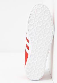 Röd sportskor med vita accenter och tre ränder. Texturerad gummisula med ett stjärnformat mönster för bättre grepp.