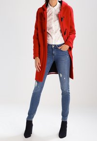 Rød uldfrakke med hætte, forlommer og toggles, båret over en hvid mønstret skjorte og blå slidte skinny jeans med sorte ankelstøvler.