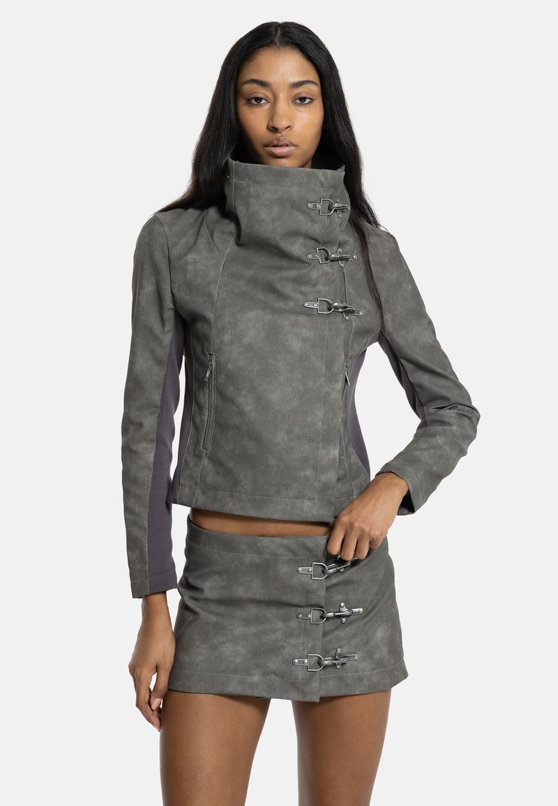 Γυναίκα ντυμένη με γκρι faux leather μπουφάν και ταιριαστή φούστα με πολλαπλές ασημένιες αγκράφες, στέκεται μπροστά σε μονόχρωμο φόντο.