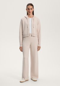 Ljusrosa kort hoodie med dragsko, parad med matchande vida byxor. Vit T-shirt under och beige sneakers syns. Mjuk textur.