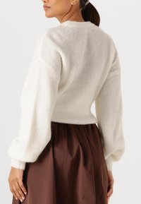 Frau mit braunen Haaren im Pferdeschwanz, die einen weißen Strickpullover und einen hoch taillierten braunen Rock trägt, von hinten und seitlich gezeigt.