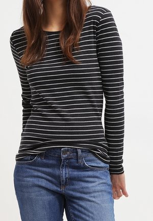 Kobieta ubrana w czarną bluzkę z długim rękawem z białymi poziomymi paskami i niebieskie dżinsy, stojąca na jednolitym tle.