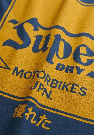 T-shirt blu con una stampa grafica gialla screpolata. Il testo recita "Super Dry Motorbikes JPN" con un distintivo effetto vintage.