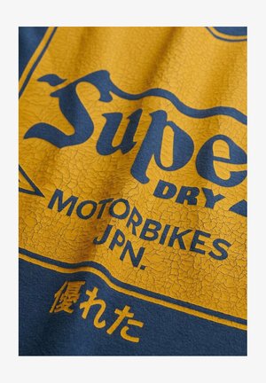 T-shirt blu con una stampa grafica gialla screpolata. Il testo recita "Super Dry Motorbikes JPN" con un distintivo effetto vintage.