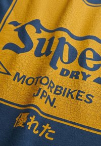 Blauw katoenen T-shirt met gescheurde gele en blauwe zeefdruktekst: "Super Dry Motorbikes JPN" met een decoratieve rand.