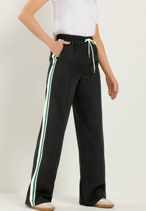 Femme portant un pantalon noir à jambes larges avec des bandes latérales blanches et vertes, un cordon blanc, et la main dans la poche, associée à un haut blanc et des baskets beiges.