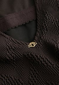 Black garment with textured, ribbed fabric and a mesh panel; features a gold logo clasp accent near the neckline and a smooth elastic band.