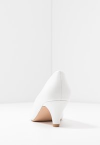 Scarpa bianca con tacco alto, punta affusolata, materiale sintetico liscio, dettagli di cucitura minimi e un tacco piccolo di colore marrone chiaro.