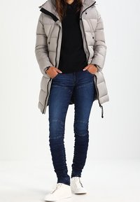 Ljusgrå puffjacka med huva, svart topp under, mörkblå smala jeans med knälappar och vita sneakers.