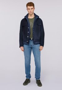 Námořnická semišová bunda se zapínáním na knoflíky a dvěma náprsními kapsami, přes zelenou košili, v kombinaci se světle modrými džínami a tmavě zelenými teniskami.