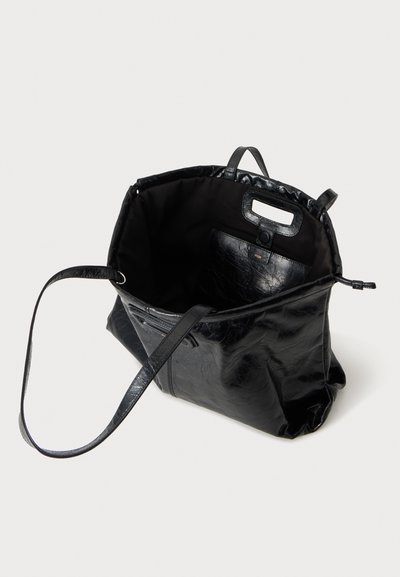 Sac cabas en cuir noir avec longues anses, ouverture sur le dessus et poche intérieure sur un fond clair uni.