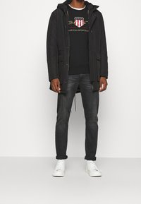 Parka negra con capucha, llevada sobre una sudadera negra con un logo rojo y blanco, combinada con vaqueros oscuros y zapatillas blancas.